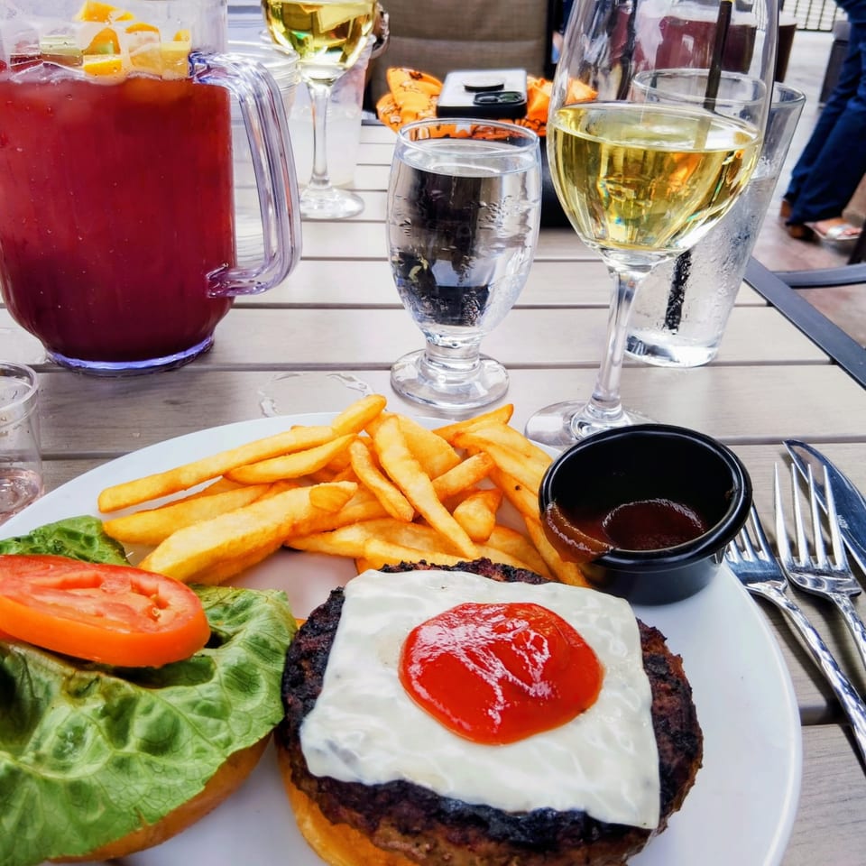 A burger with cheese and ketchup, fries, and drinks on a wooden outdoor table.