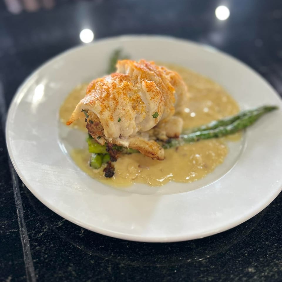 A white plate holds a stuffed lobster tail with asparagus and a creamy sauce.