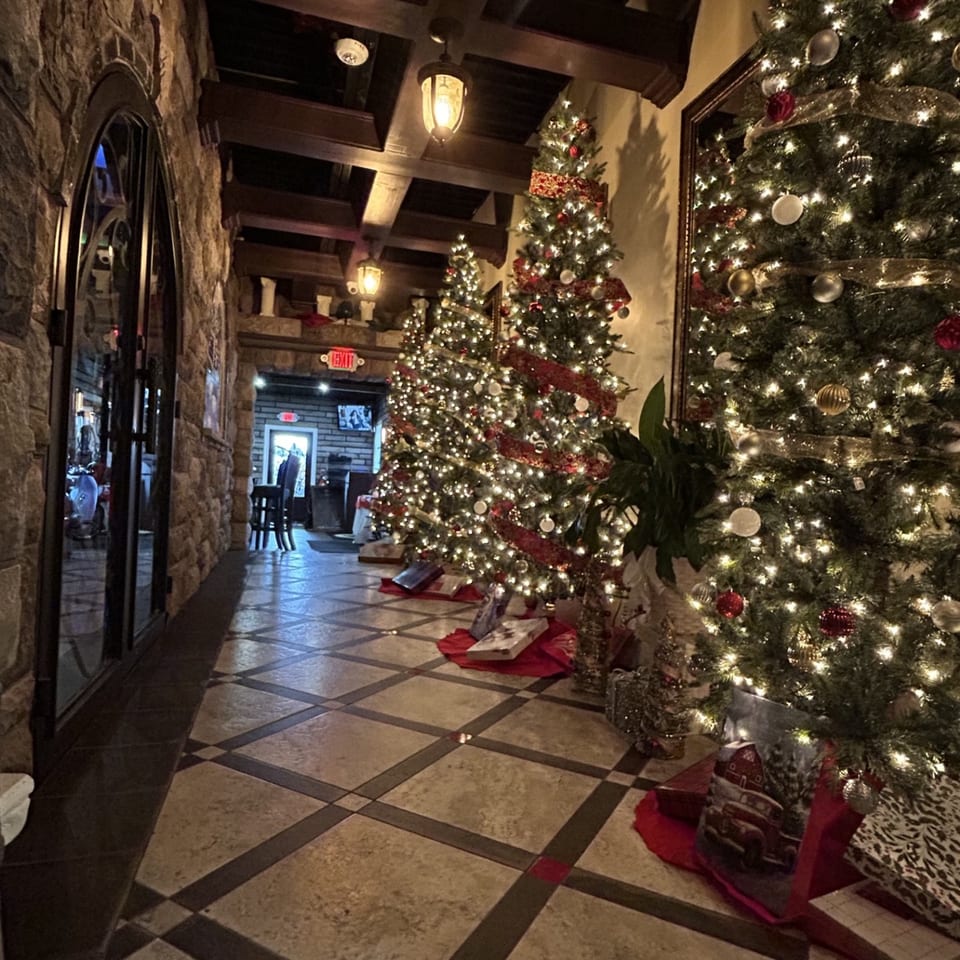 Restaurant lobby decorated with several illuminated Christmas trees, red and silver ornaments, wrapped gifts, stone walls, and tiled floors.