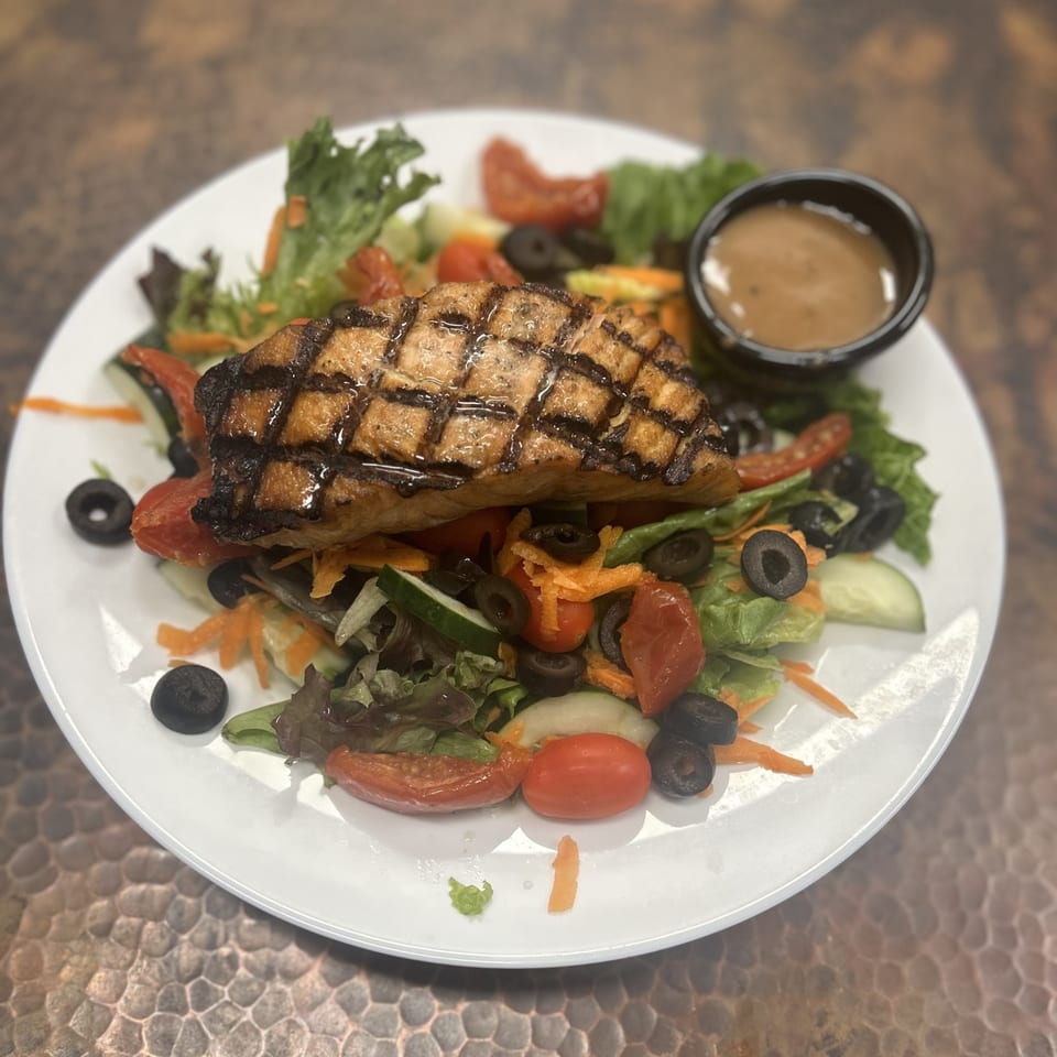 Grilled salmon salad with mixed greens, cherry tomatoes, olives, and dressing on a white plate.