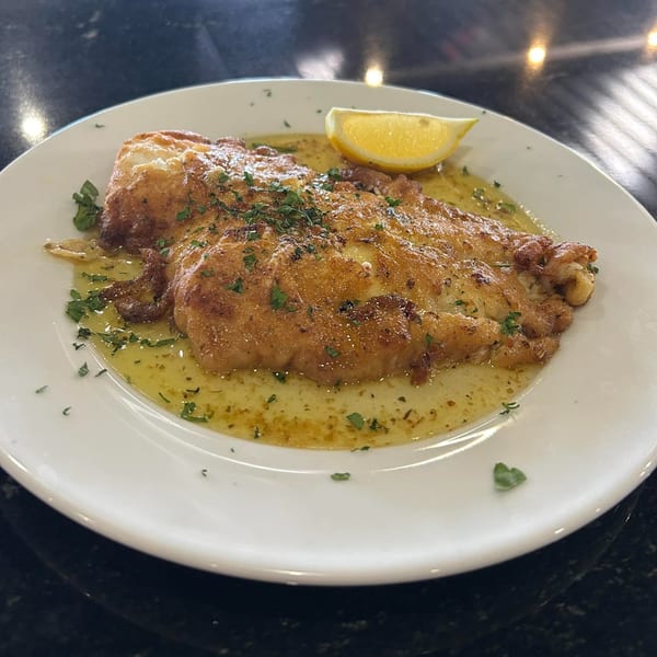 A golden-brown chicken cutlet, possibly Chicken Francese, in a lemon-butter sauce with parsley and a lemon wedge.